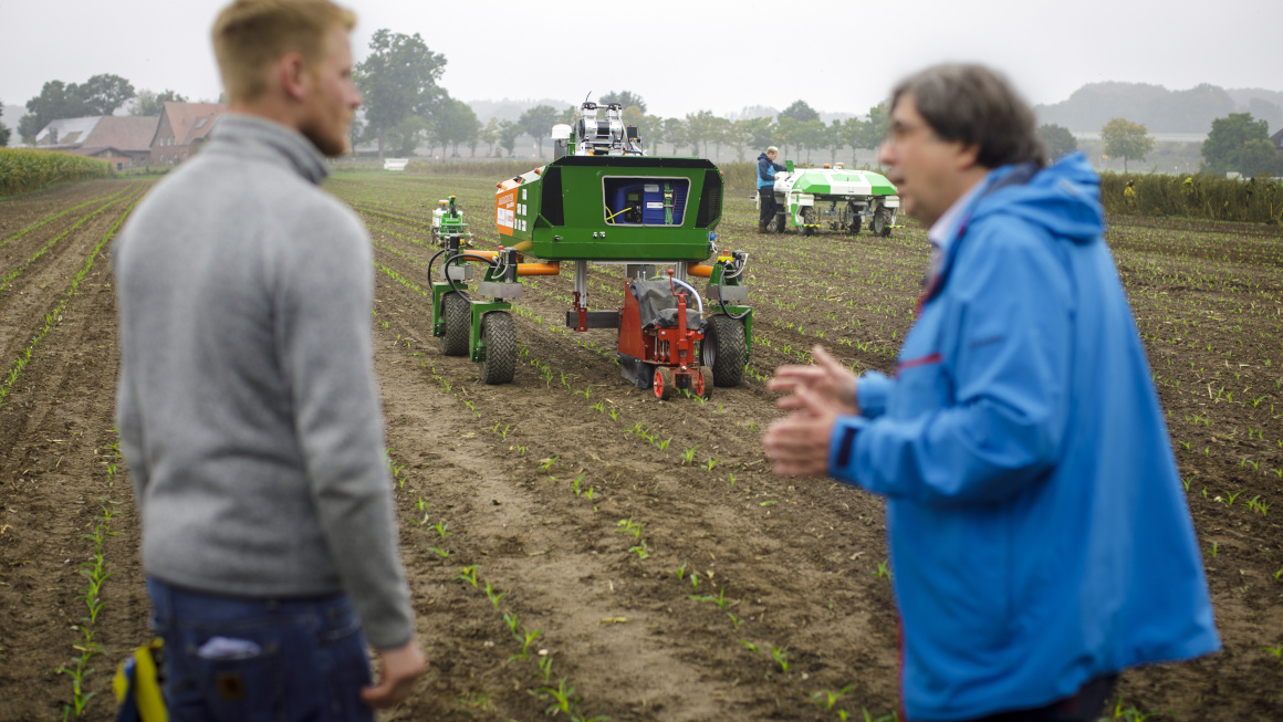 Im Vordergrund zweit Wissenschaftler im Gespräch, dahinter zwei landwirtschafltiche Roboter für die Feldarbeit im Einsatz