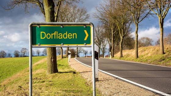 Das Straßenschild "Dorfladen" ist vor einer Landstraße und einem Feld zu sehen.