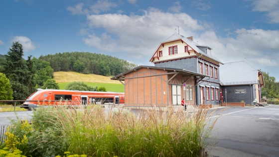 sanierter Bahnhof im ländlichen Raum mit einem Zug, im Hintergrund ein Hügel mit einem Feld und Wald