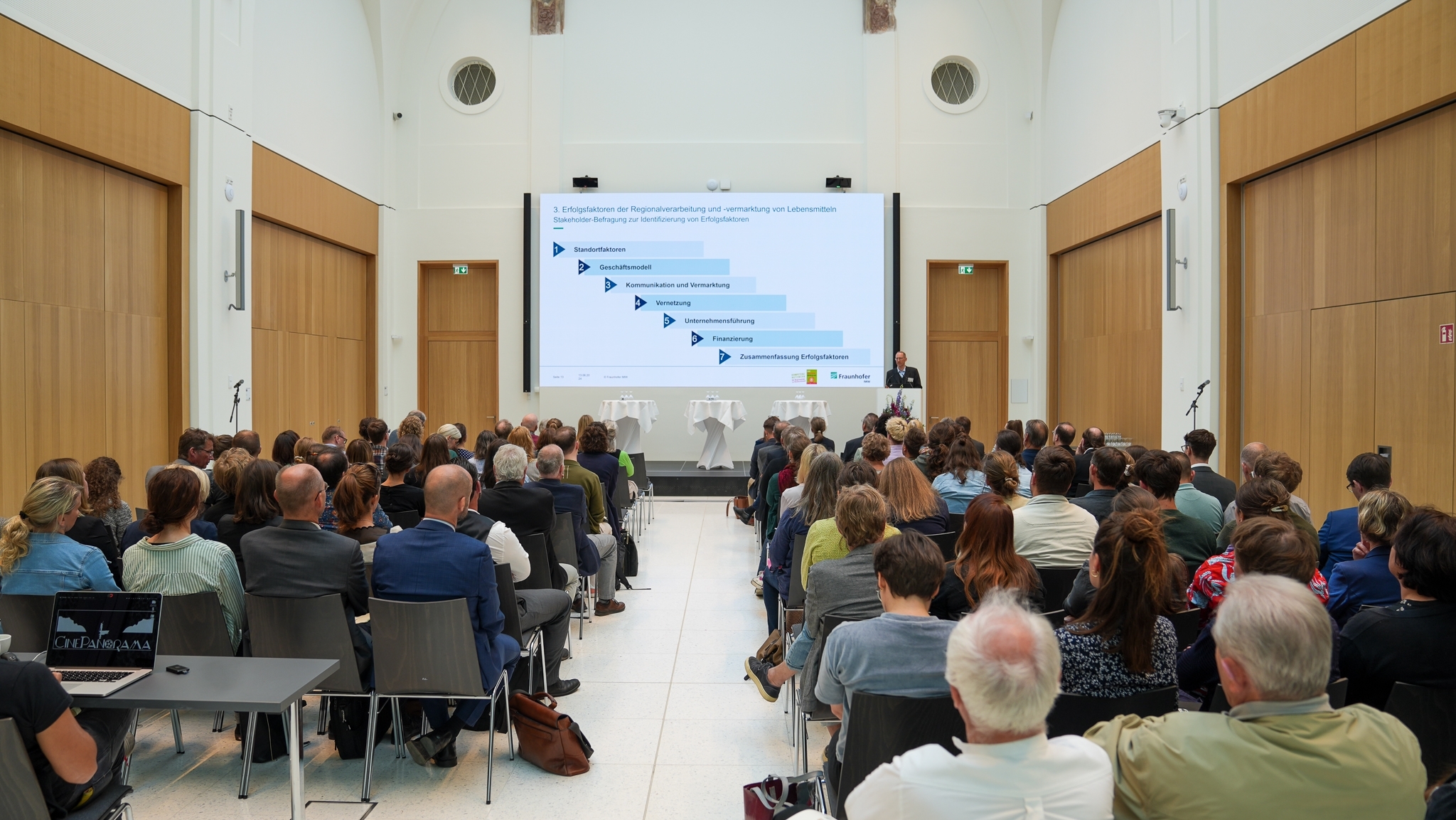 Ein Fachkongress zum Thema "Erfolgreiche Regionalvermarktung". Viele Leute schauen gespannt nach vorne. Ein Mann hält eine Präsentation auf einem Podium. Er präsentiert Erfolgsfaktoren.