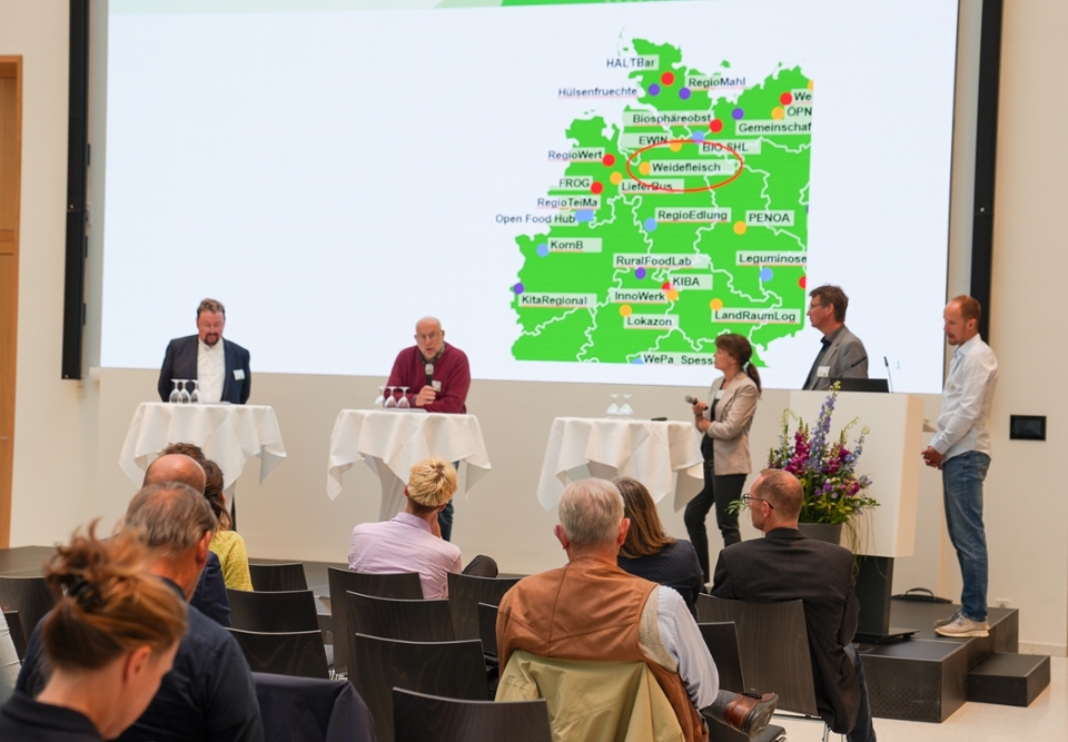 Ein Fachkongress zum Thema "Erfolgreiche Regionalvermarktung". 5 Personen stehen auf der Bühne und stellen beispielhaft Projekte der Regionalvermarktung vor. Im Hintergrund ist eine Landkarte zu sehen.