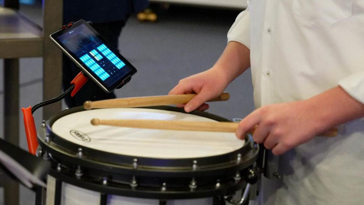 Ein Tablet mit einen Musikprogramm steckt in einer Halterung - daneben spielt eine Person auf der Trommel.