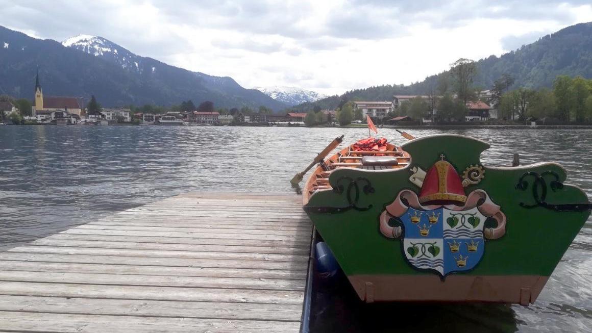 Am Steg liegt ein Ruderboot mit Wappen am Heck, im Hintergrund der Tegernsee und die Alpen