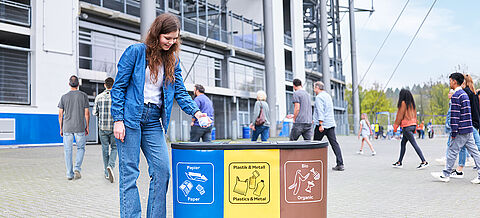 Eine Frau wirft ihren Abfall in einen Behälter vor einem Fußballstadion. Der Behälter ist mit den Aufschriften 'Papier', 'Plastik & Metall' sowie 'Bio' gekennzeichnet.