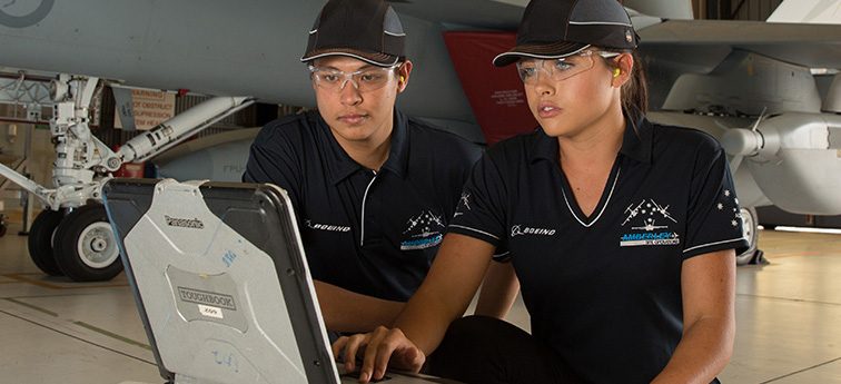 Two people in bump caps, safety glasses and ear protection working on a laptop