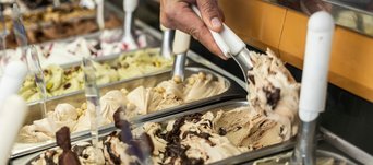 Eine Hand schöpft mit einem Spatel Eis aus einer Eistheke mit verschiedenen Sorten in Metallbehältern.