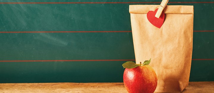 Foto von einem Apfel und einer Papiertüte mit Wäschklammer und einem daran befestigten Herzen auf einem Schultisch vor einer  Tafel