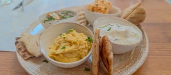 Auf einem Holztisch steht ein heller Teller, darauf kleine Schüsseln mit Auberginensalat, Humus, Fava und Tzatziki sowie dreieckige Pitabrot-Stücke. 