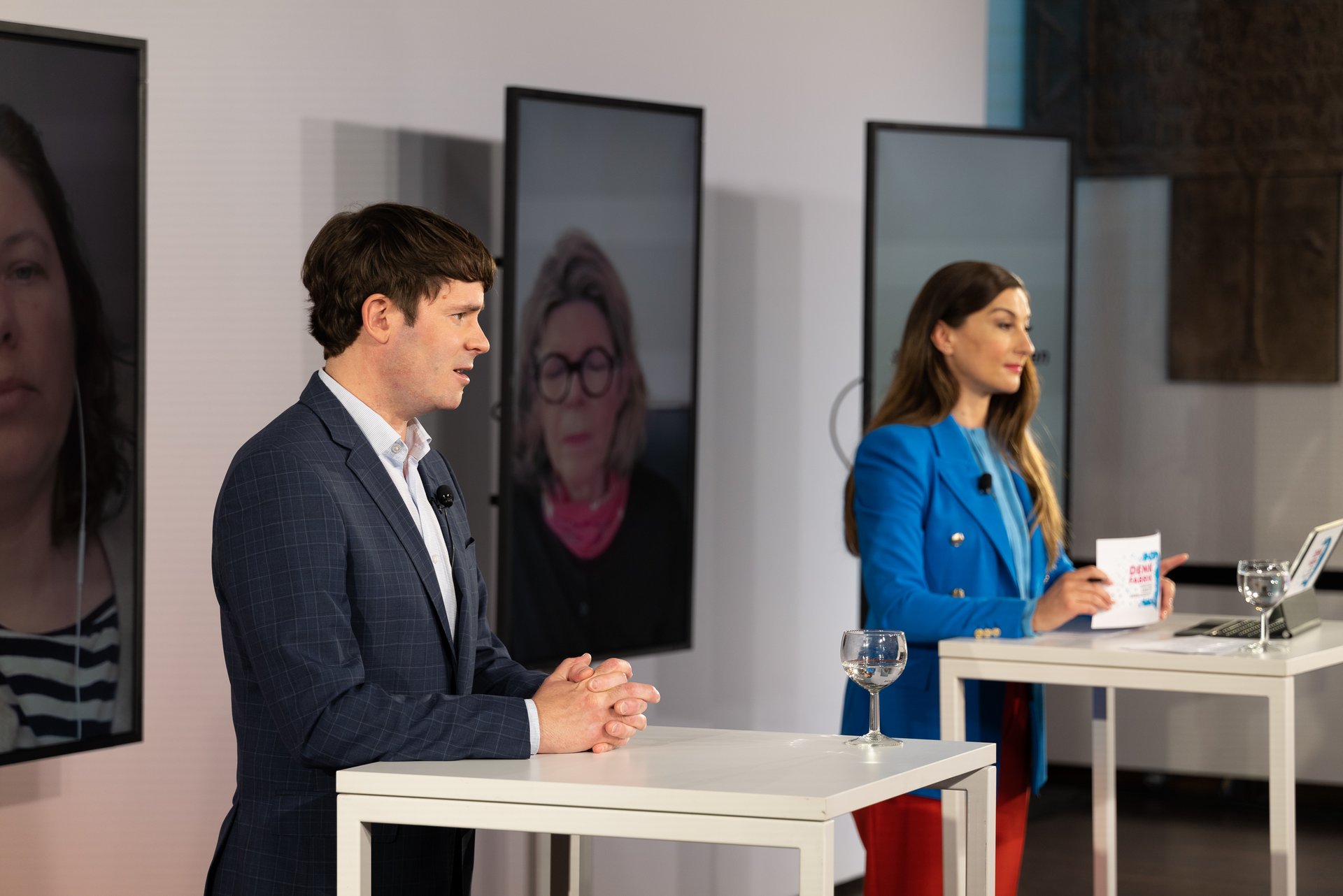 Ein Mann und eine Frau stehen an Stehtischen auf der Bühne vor mehreren Monitoren mit zugeschalteten Personen.