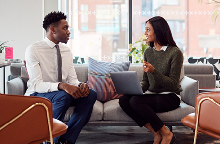 Woman on her laptop talking with man. Both sitting on couch looking at each other.