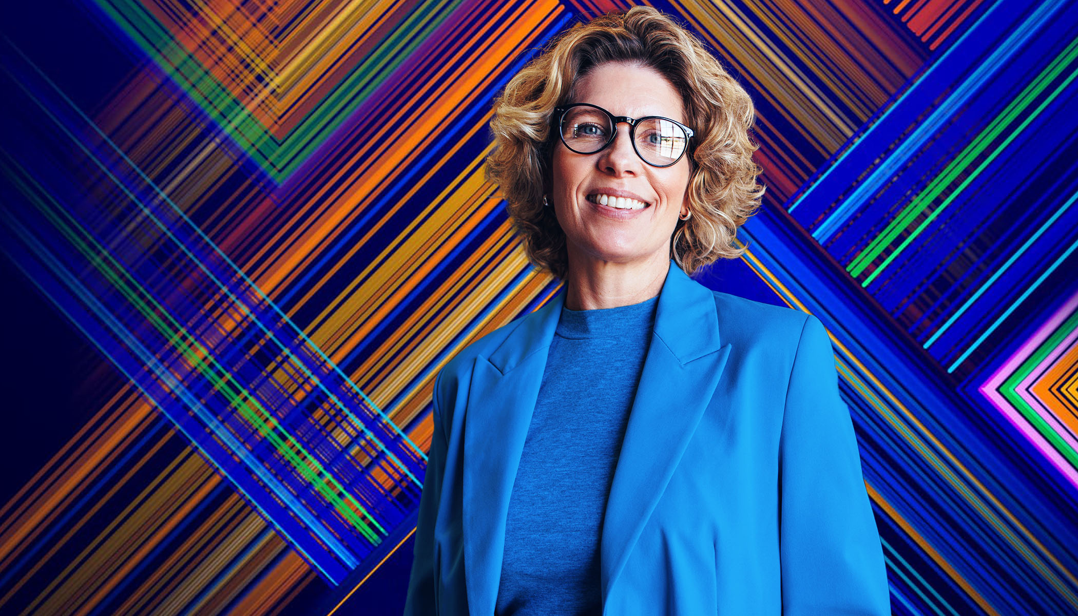 A woman wearing glasses and a blue blazer stands in front of a background of colorful lines