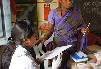 A woman dressed in a purple sari returns her self-collection vaginal swab to a clinic worker, dressed in medical whites, for HPV