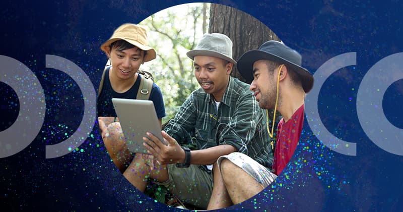 Three people sit near a large tree and smile as they look at a tablet one of them is holding