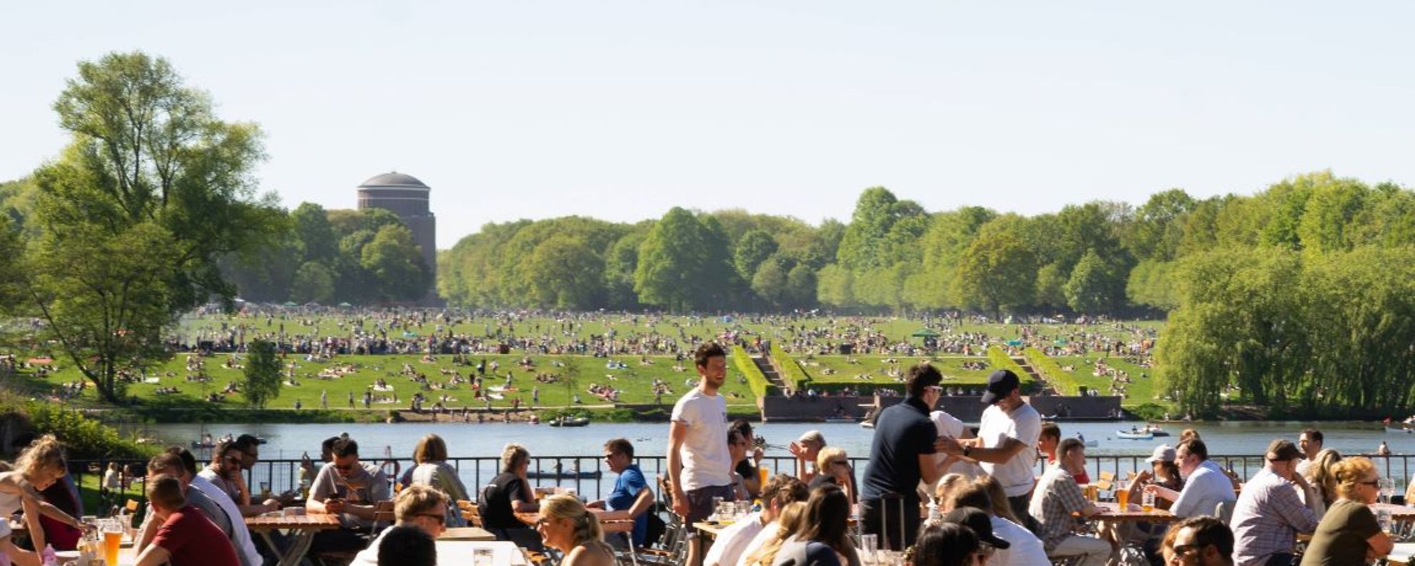 Sommer im Hamburger Stadtpark, im Vordergrund sitzen Menschen an Bierzeltgarnituren, im Hintergrund ist das Planetarium zu sehen.