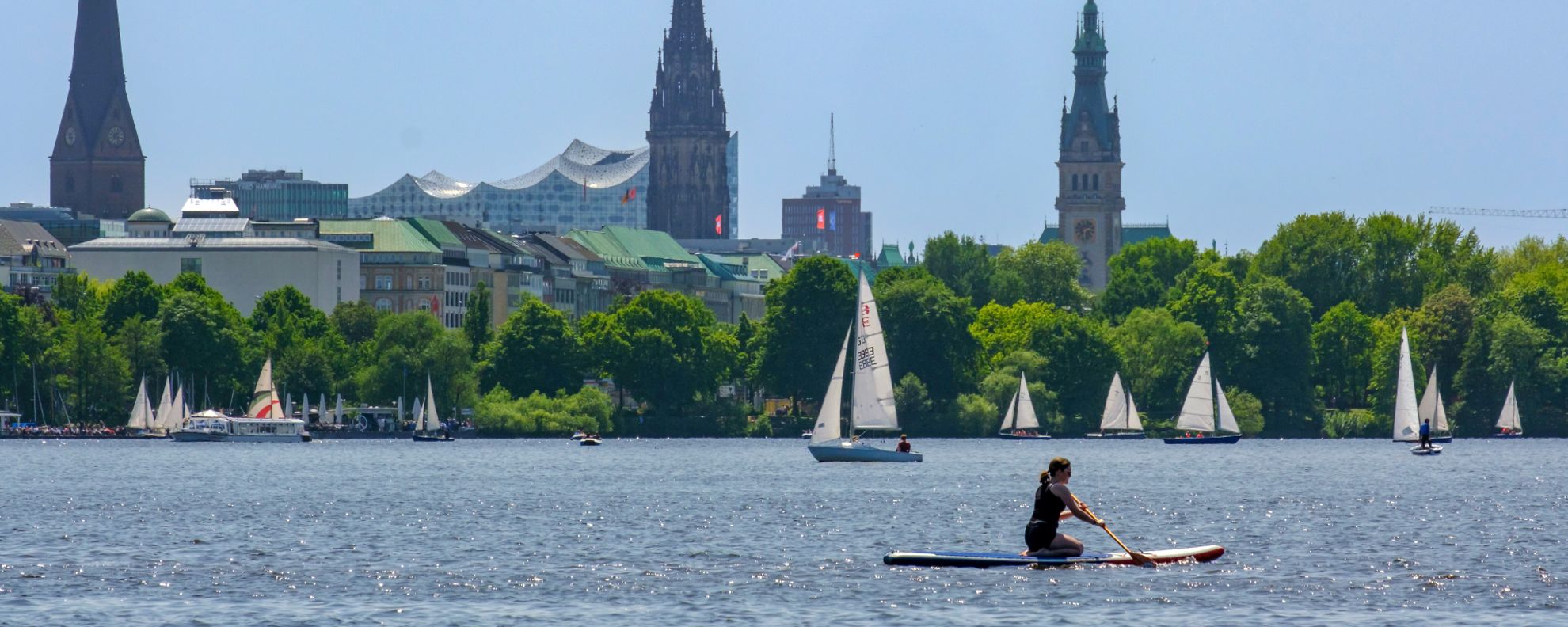 Auf dem Wasser paddelt eine Frau und es fahren mehrere Segelboote. Im Hintergrund sieht man das Stadtpanorama Hamburgs.