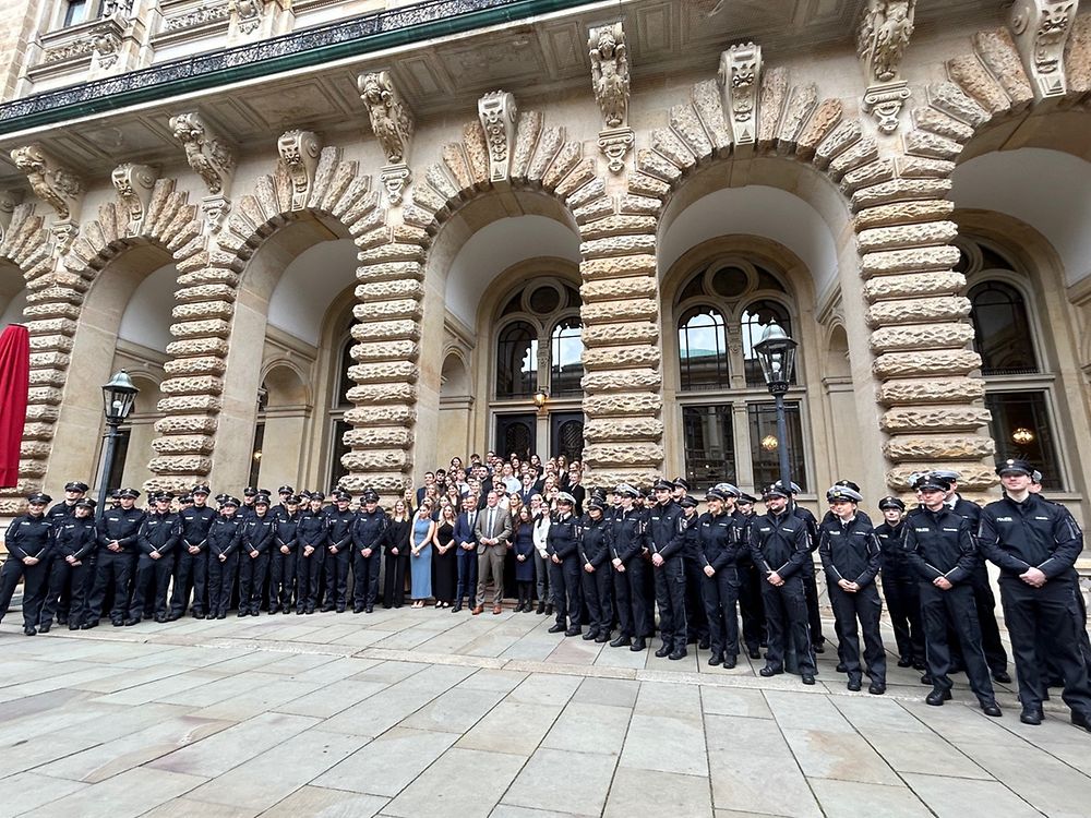 Polizisten und Polizistinnen stehen in Uniform nebeneinander. In der Mitte der Gruppe stehen Personen in Kleidern sowie Anzügen.