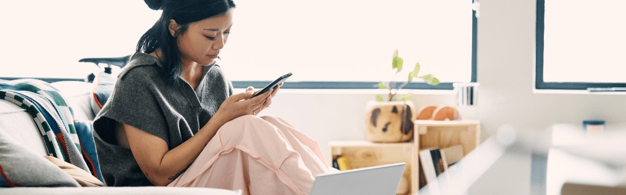 A woman sitting on the couch holding a smartphone with an HP Envy laptop near her.