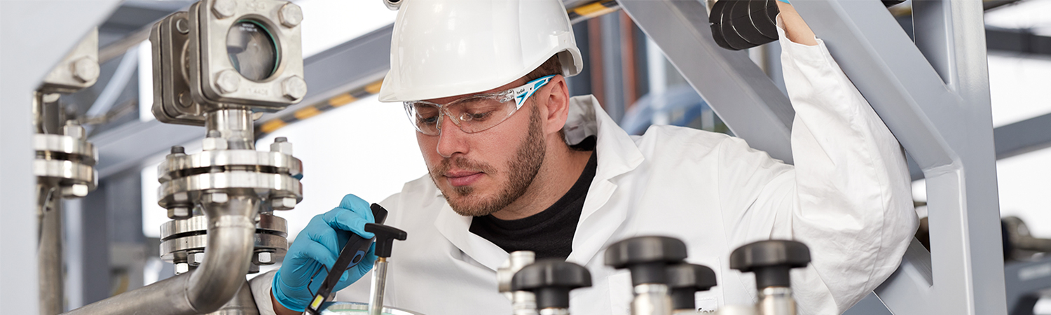 Mann mit Laborkittel, Sicherheitsbrille und Schutzhelm in einem Technikum für biobasierte Produkte, der an einer Maschine eine Messung vornimmt