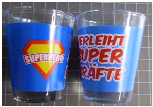 Abgebildet sind zwei Schapspinnchen mit buntem Aufdruck in blau-gelb-rot und der Aufschrift Superhero verleiht Superkräfte