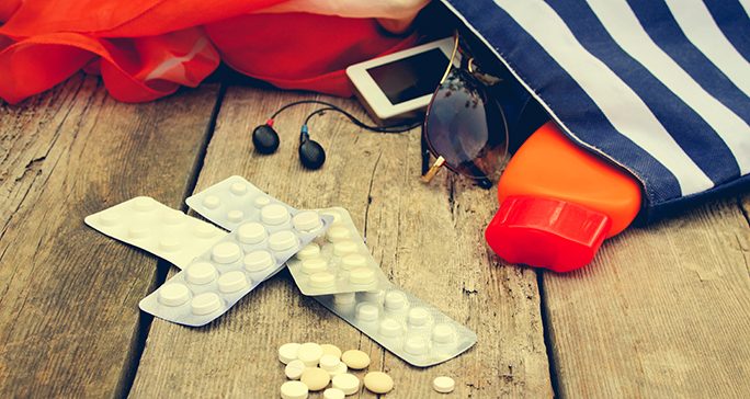 Colorful travel tote on wooden floor spilling out pills, earbuds, sunscreen, sunglasses and other personal items