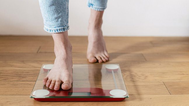 Person wearing blue jeans takes a step onto a clear digital scale on a hardwood floor.