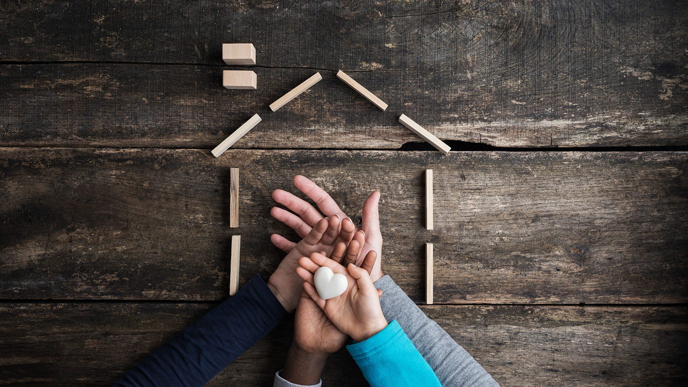 Close-up of 4 stacked adult and kids' hands, palm up, outlined by a house, with a stone heart in the middle