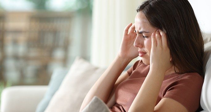 A grimacing woman sits on a couch with her hands on her temples.