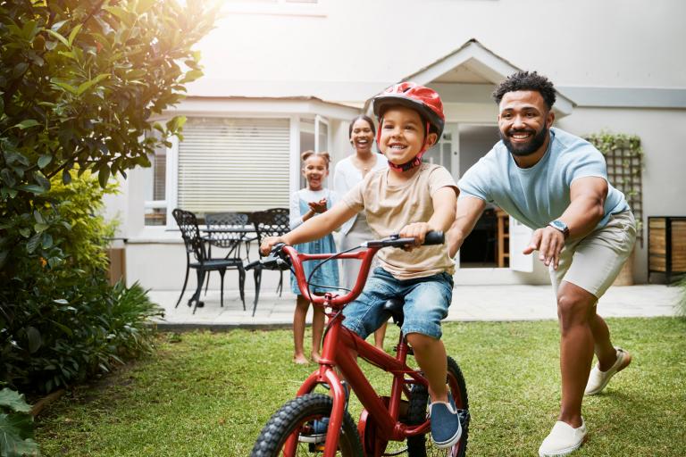 A man pushes his son on a bicycle in their front yard as his wife and daughter watch.