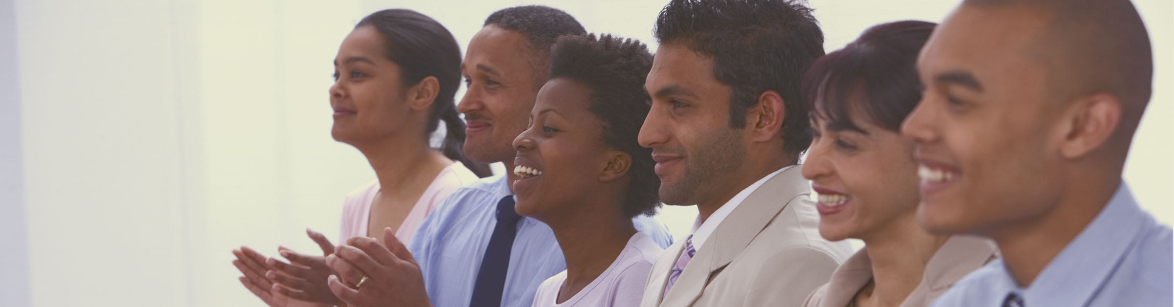 A smiling group of people of different genders, ages, races and ethnicities, seated at a conference table clapping their hands