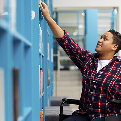 A person with brown skin wearing a plaid shirt and sitting in a wheelchair reaches up to take a book off a blue bookshelf