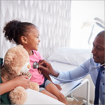 With her mother beside her, a young, Black child smiles as a male, Black doctor listens to her lungs with a stethoscope