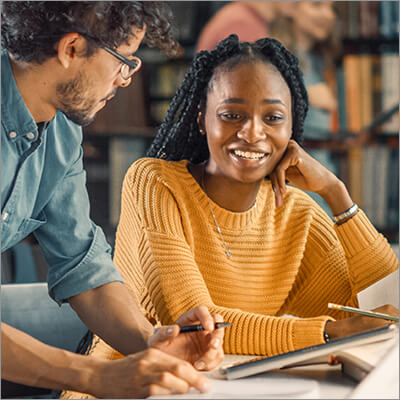 Two college students, a woman and man, discuss the books and papers on a table in front of them. Behind them, two students walk past library shelves