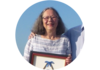 A woman with medium length grey hair wearing a striped shirt, standing outside, holding an award.