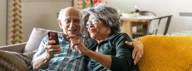 deux personnes âgées souriantes regardent leur mobile dans leur canapé