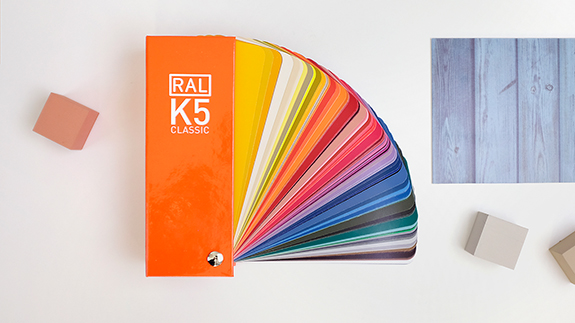 Der RAL K5 Farbfächer gloss liegt aufgefächert auf einem weißen Untergrund. Daneben liegen 3 jeweils einfarbige Würfel verteilt sowie oben rechts ein Stück Holz. | RAL FARBEN

The RAL K5 Colour fan gloss lies fanned out on a white background. Next to it are 3 single-coloured cubes and a piece of wood at the top right. | RAL COLOURS