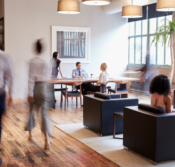 A group of people sitting at a table in an office, some engrossed in conversation while others are focused on their laptops.