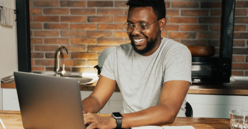 A man  wearing glasses, gazes at his laptop screen with. He is at his home's kitchen table.