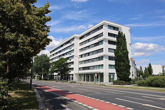 Foto des weißen Sozialministeriums. Davor eine leere Straße, Bäume und blauer HImmel