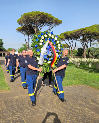 Mehrere THW-Kräfte in blauer Einsatzkleidung gehen auf einem Weg eines Friedhofs. Die vorderen beiden Personen tragen ein Blumengesteck mit blauen und weißen Blumen und Deutschlandflagge.