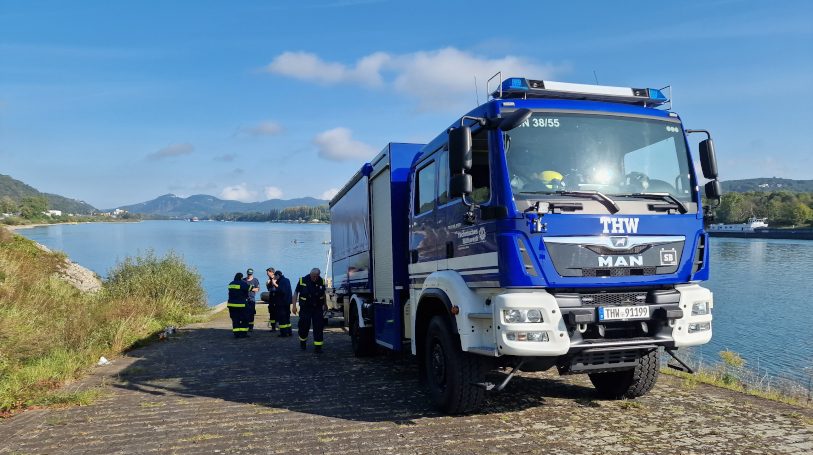 An einem Fluss steht ein blaues Einsatzfahrzeug des Technischen Hilfswerk. An der Seite und Rückseite des Fahrzeugs sind mehrere Kräfte in blauer THW-Kleidung beschäftigt.