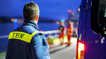 Ein THW-Helfer in blauer Einsatzkleidung mit THW Beschriftung steht neben einem blauen Einsatzfahrzeug. 