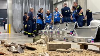 Eine Gruppe von Menschen, teils in blauen Westen, teils in blauer THW-Einsatzkleidung, steht zusammen. Vor ihnen stehen silberne Metallkisten, die geöffnet sind. Davor wiederum befindet sich eine Fläche mit Sand und Kulturgütern.