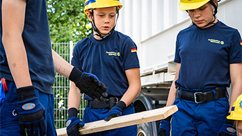 Auf dem Bild sind drei Personen, alle in blauer THW-Kleidung. Der Junge in der Mitte hält eine Holzplanke.