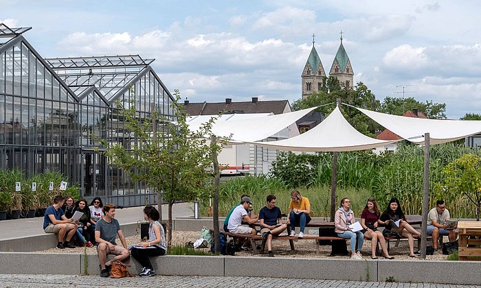 Studierende des TUM Campus Straubing für Biotechnologie für Nachhaltigkeit sitzen vor Gewächsgärten in Straubing, im Hintergrund die Kirche St. Peter und das Technologie- und Förderzentrum. 