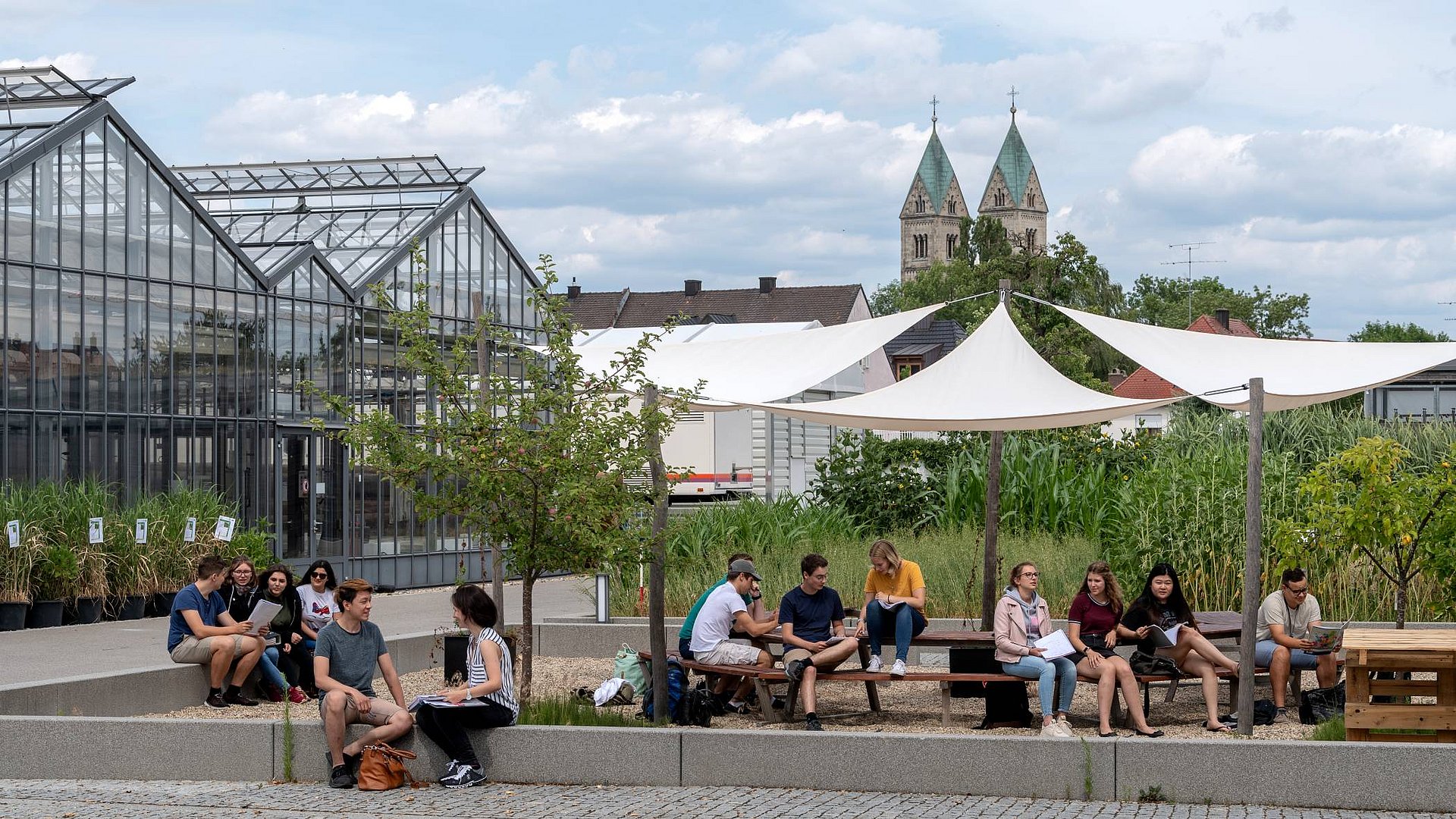 Studierende des TUM Campus Straubing für Biotechnologie für Nachhaltigkeit sitzen vor Gewächsgärten in Straubing, im Hintergrund die Kirche St. Peter und das Technologie- und Förderzentrum. 