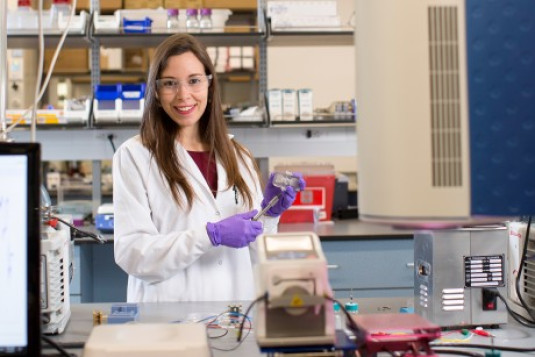 Daniela Blanco, una joven latina de cabello castaño, viste una bata de laboratorio blanca, gafas de seguridad y guantes de plástico entre equipos de química en un laboratorio bien equipado. Pausa su trabajo con unos instrumentos para sonreír a la cámara.