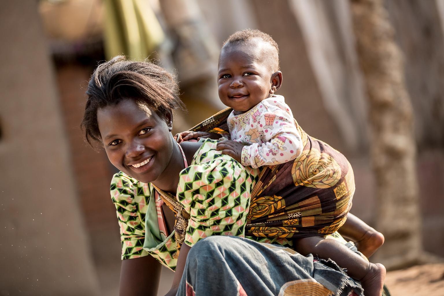 An African woman carrying her baby on her back, both smiling for the camera