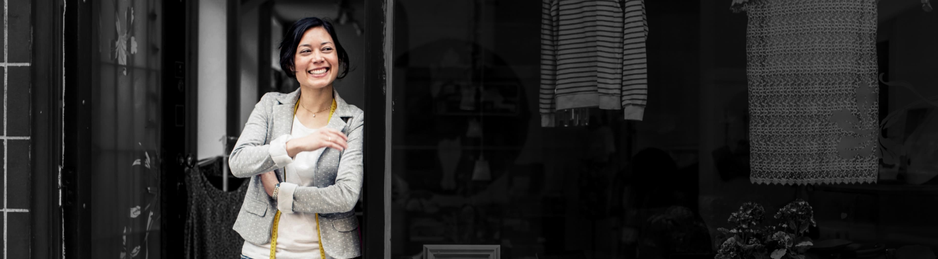 A woman in front of her alterations business smiling and holding a measuring tape